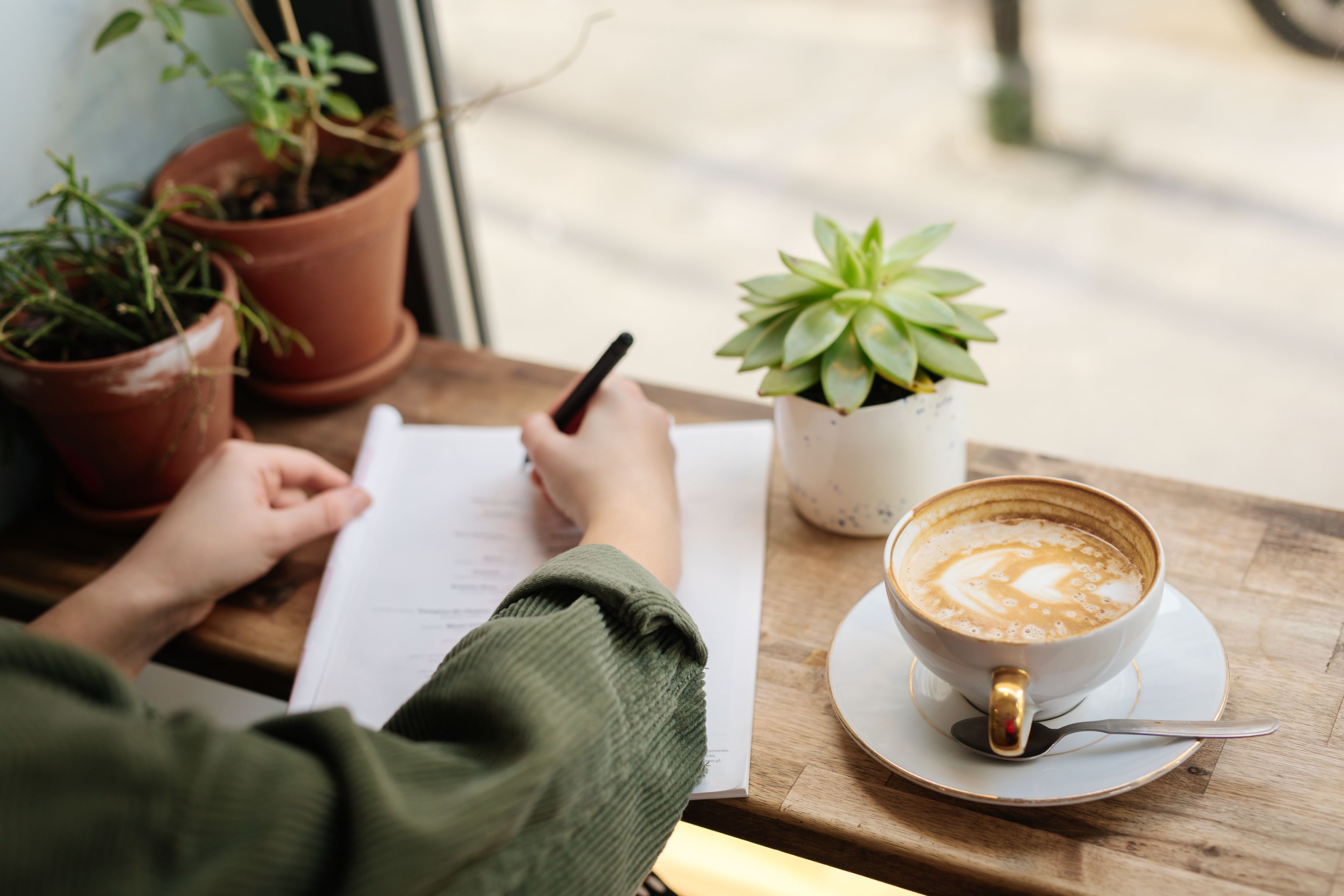 Person skriver på papirer med kaffekopp og planter på bordet